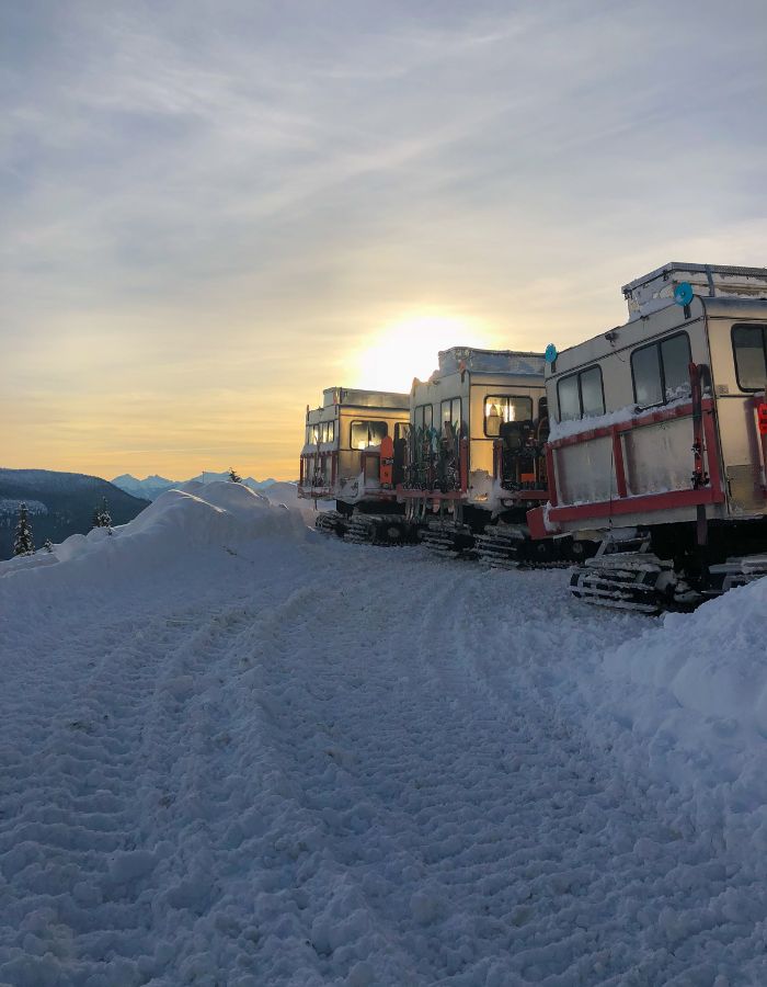 View looking at the back of 3 snow cat vehicles (used to transport people over deep snow) parked at the top of a mountain.