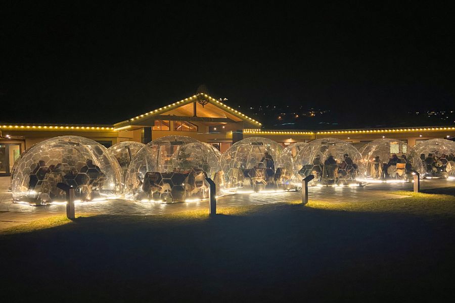 The same patio at night in the winter. Clear domes with tables inside adorn the patio. The domes are lit up and there are small white lights along the top of the building. 