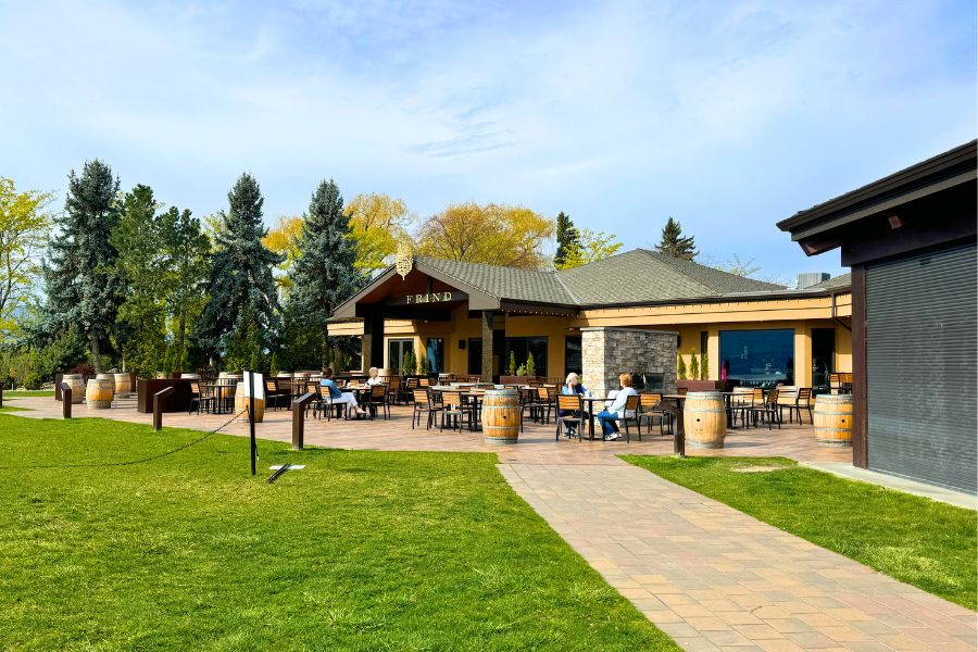An outside patio with tables and wine barrels surrounded by grass. A building stands in the background with the words "Frind" written on it.
