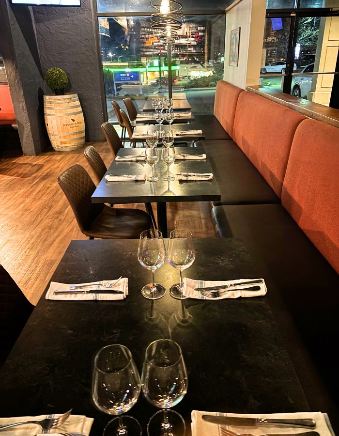 View of tables side by side - a booth on one side and chairs on the other side. The tables are black with wine glasses and cutlery on top.