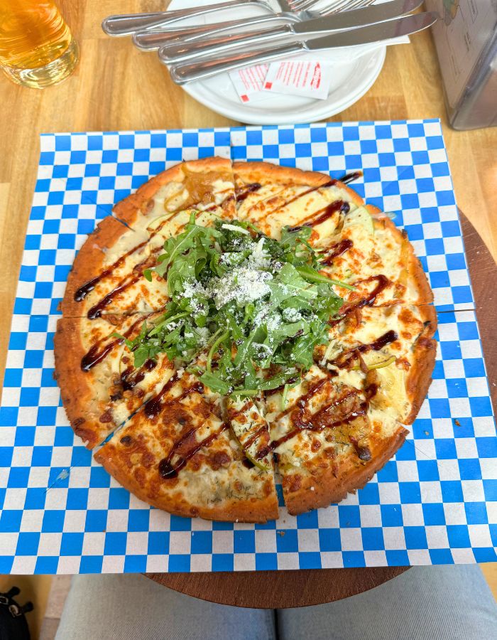 A pizza on a piece of blue and white checkered parchment paper. The pizza has cheese, apples and greens on the top with a white sauce.