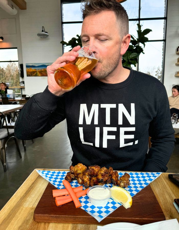 Man wearing a black sweatshirt that says "MTN LIFE" on the front. He is drinking a beer and has a plate of chicken wings in front of him