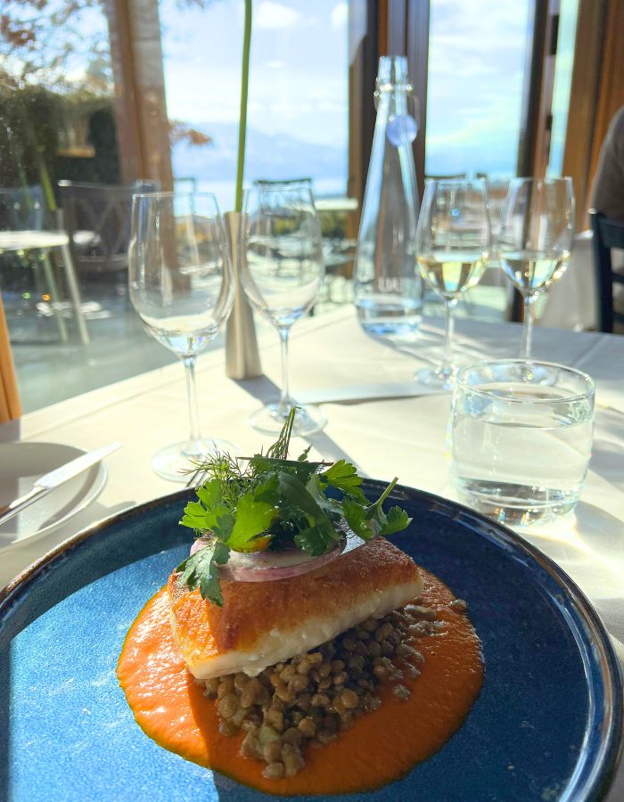 Fish on a blue plate of lentils topped with greens. Wine glasses are on the table and a faint lake view is in the distance